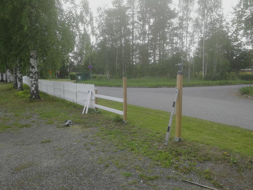 Kuva esittää valkoista aitaa, jonka osia on vielä rakentamatta. Aidan vieressä on puita ja taustalla näkyy tie sekä metsää. Valo on pehmeää ja luonnonläheistä.