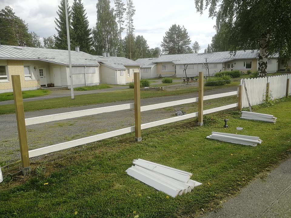 Maisema, jossa on valkoinen aita rakenteilla, ruskeat pylväät, valkoiset laudat maassa, vihreä ruoho ja rakennuksia taustalla, valoisa sää.