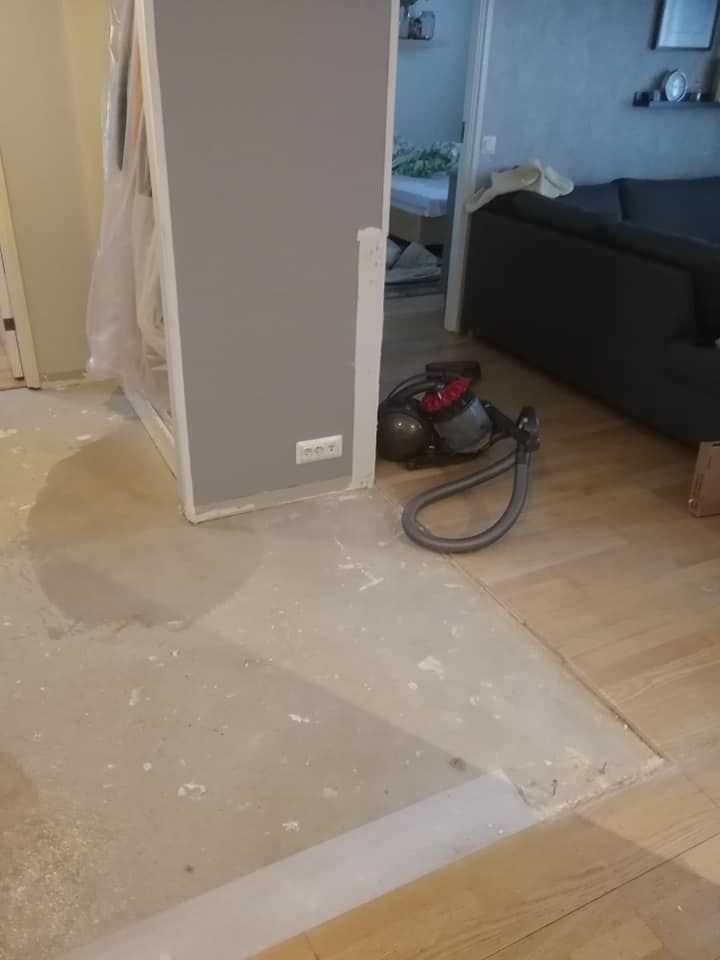 Huoneessa on harmaa seinä, pölynimuri ja osittain asennettu vaalea puulattia. Lattian keskellä on sementtipohja.