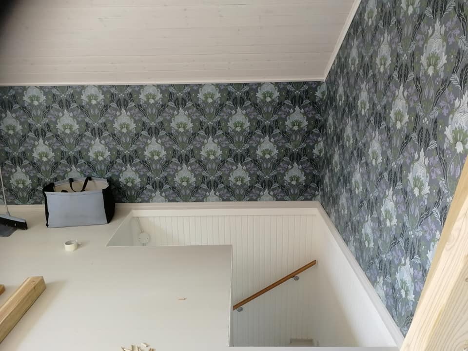 Huoneessa on tapetti, jossa on kukka-aiheinen kuviointi, valkoinen katto ja portaat. Lattialla on työvälineitä ja säilytyskassi.