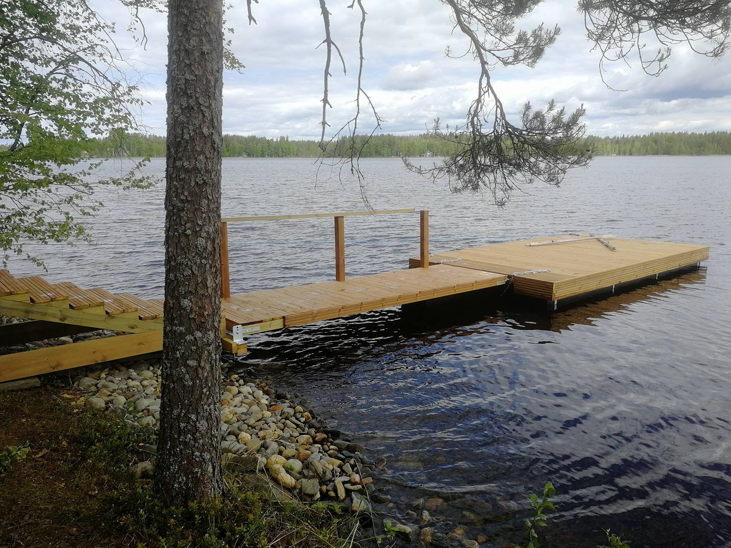 Laituri järvellä, puinen rakenne, kivikkoinen ranta, puu edessä, pilvinen taivas.
