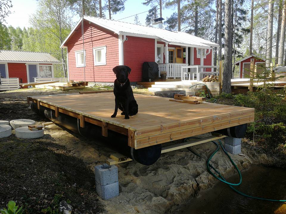 Ulkona on aurinkoinen päivä, ja musta labradorinnoutaja istuu puisen kelluvan laiturin päällä. Taustalla näkyy punainen talo valkoisilla ikkunankehyksillä ja katolla, sekä muita rakennuksia ja puita.