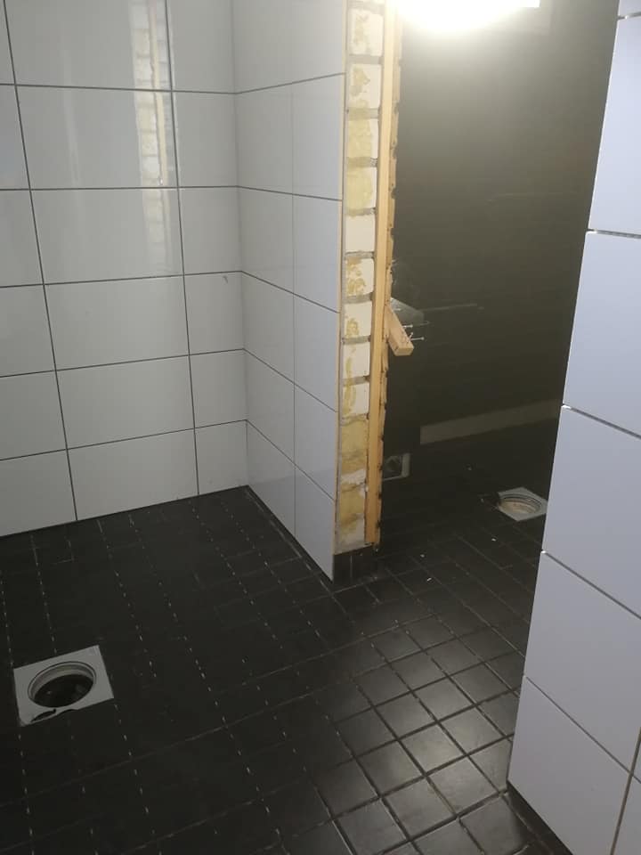 Kylpyhuoneen sisätila, jossa on musta laattalattia, valkoiset seinälaatat ja avoin seinärakenne, jossa näkyy puu ja eristys.