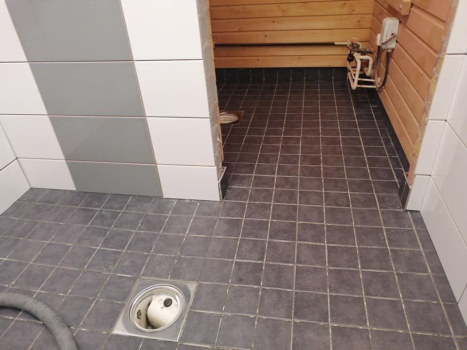 Kuva kylpyhuoneesta, jossa on harmaat lattialaatat, valko-harmaat seinälaatat ja puinen sauna. Lattialla on metallinen viemäri ja harmaa putki. Saunan seinällä on putkisto ja valkoinen sähkölaatikko.