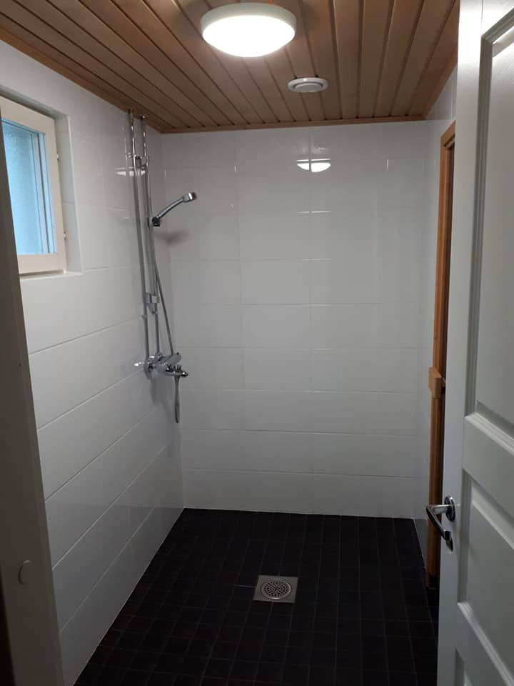 Kylpyhuoneessa on valkoiset seinälaatat, tumma lattia, puinen katto ja suihku.