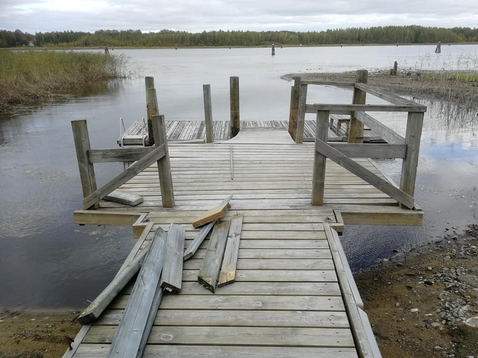 Harmaa puinen laituri järvellä, jossa on keskeneräisiä kaiteita ja puutavaraa, pilvisen sään alla.