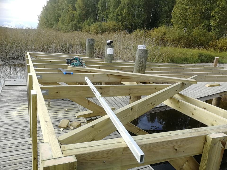 Kuva esittää puisen laiturin rakennusvaihetta, jossa näkyy runko, työkaluja ja taustalla luontoa.