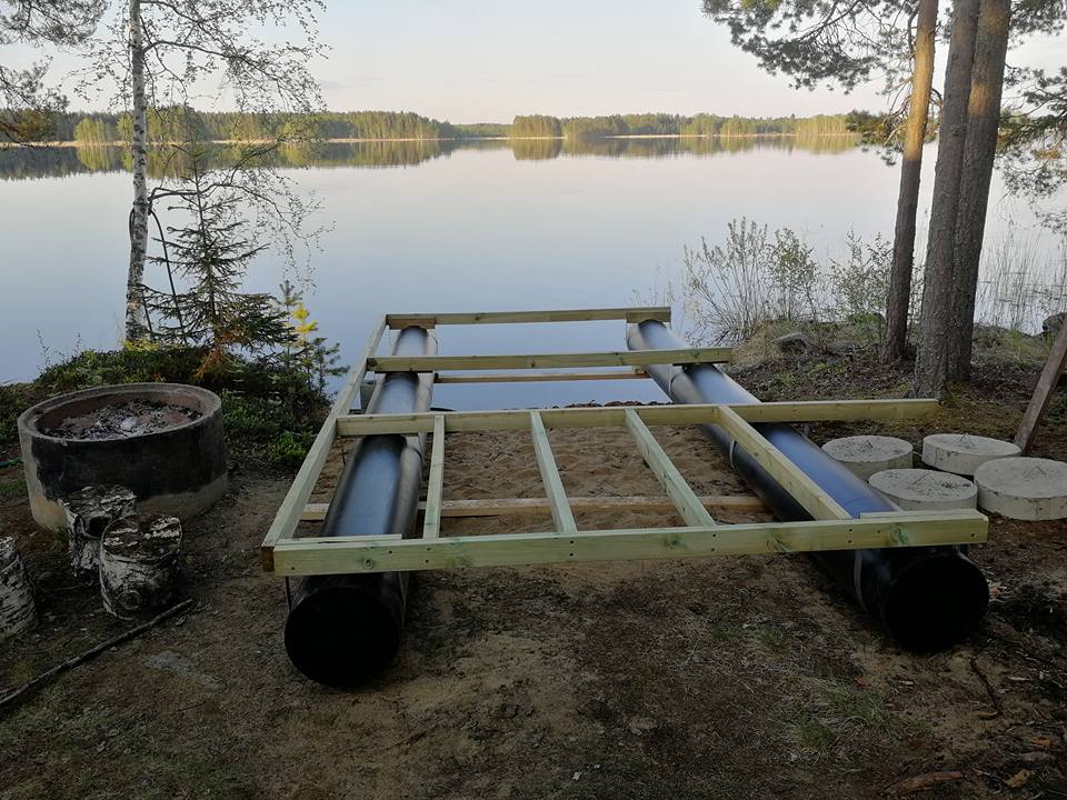 Järven rannalla on rakenteilla oleva lautta, jossa on mustat kellukkeet ja puinen runko. Taustalla näkyy tyyni järvi ja metsäinen maisema.