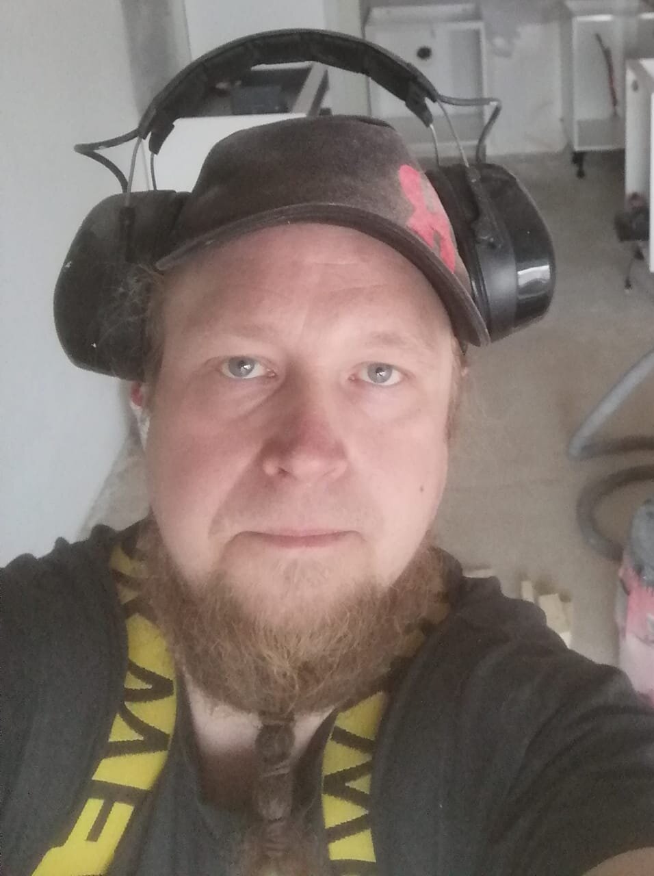 Lähikuva miehestä, jolla on parta, hattu ja kuulosuojaimet, sisätiloissa, jossa on valkoisia kaappeja.