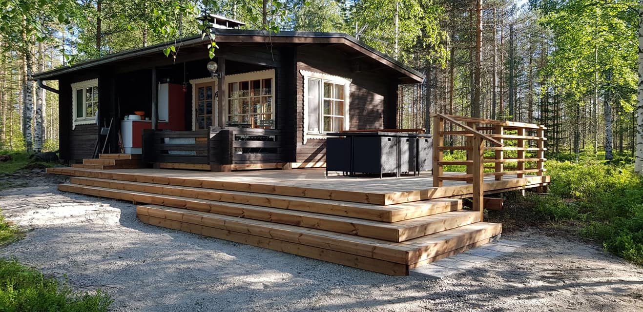 Metsämökki, jossa on puinen terassi ja portaat, sijaitsee metsäisessä ympäristössä aurinkoisena päivänä.
