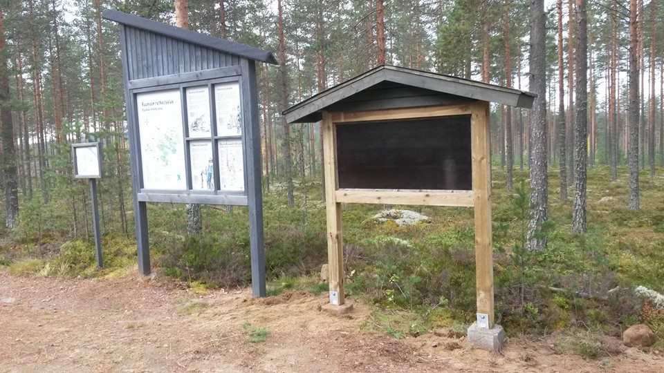 Metsämaisemassa on kaksi opastaulua, joista toisessa on karttoja ja toinen on tyhjä musta taulu. Taustalla näkyy tiheä havumetsä.