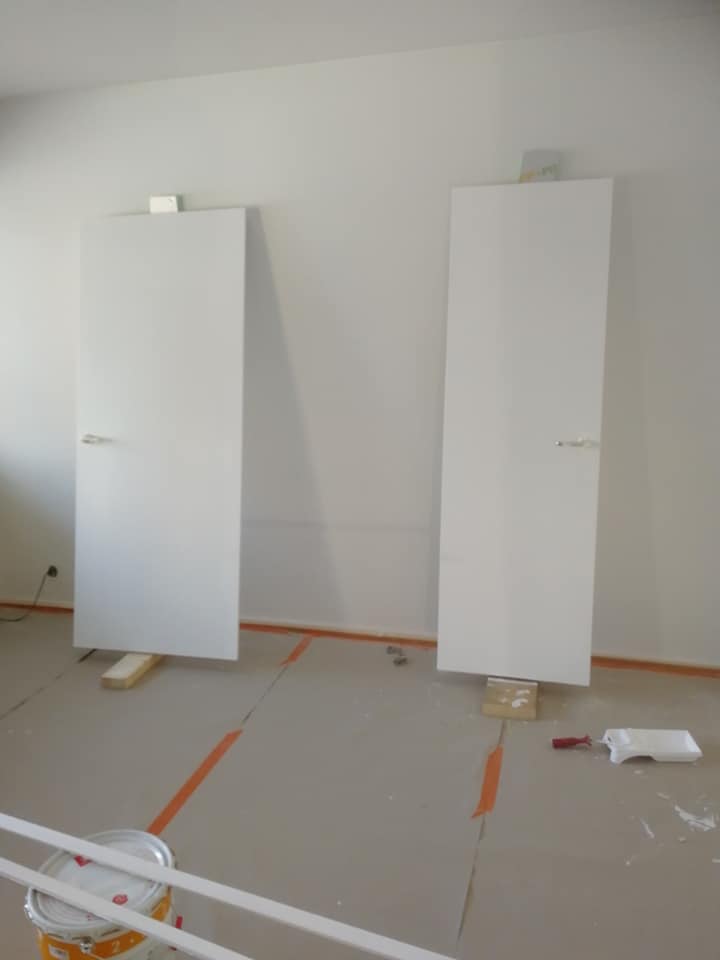 Kaksi valkoista ovea nojaavat seinää vasten, maalipurkki ja maalarintarvikkeita lattialla, oranssi teippi lattialla.