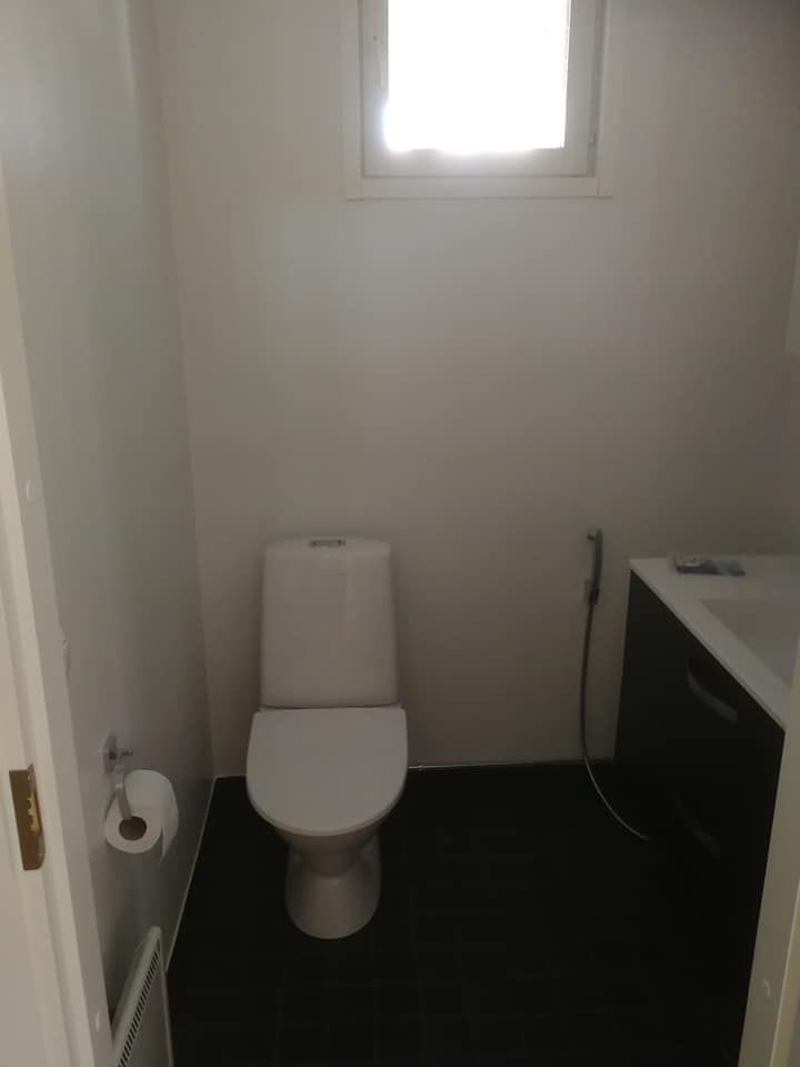 Pieni, valkoinen kylpyhuone, jossa on wc-istuin, pesuallas ja pieni ikkuna. Lattialaatat ovat tummat.