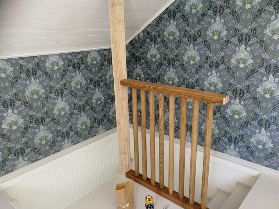 Kuva näyttää sisätiloissa olevan portaikon, jossa on puinen kaide ja pylväs. Seinät on tapetoitu kuviollisella tapetilla, jossa on harmaita, vihreitä ja valkoisia sävyjä.