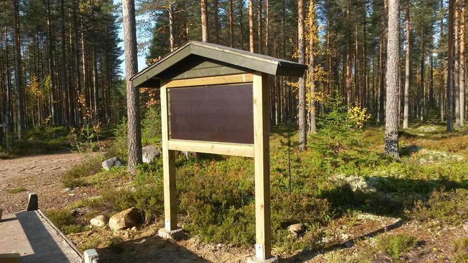 Metsämaisemassa on puinen infotaulu, jossa on tumma tausta ja vihreä katto. Taustalla näkyy korkeita puita ja maassa on sammalta ja pensaita. Valo on kirkas ja aurinkoinen.