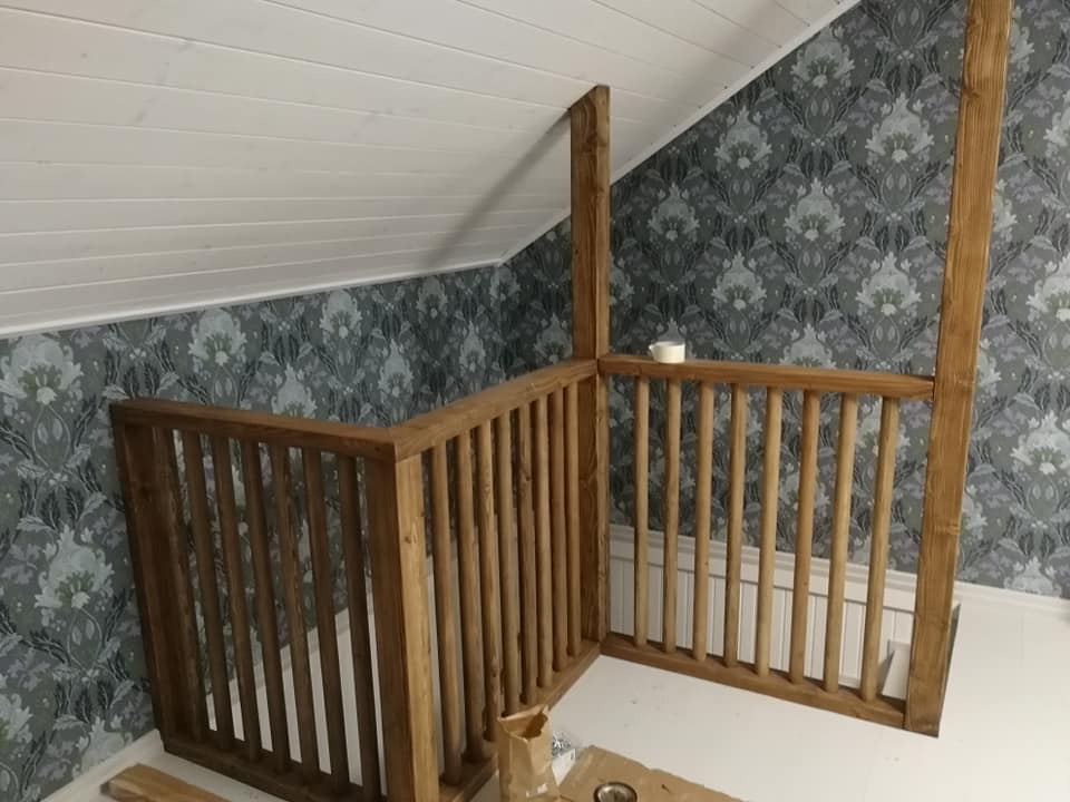 Puinen kaide, jossa on pystysuorat säleet, seisoo huoneessa, jossa on kuvioitu tapetti ja valkoinen katto.