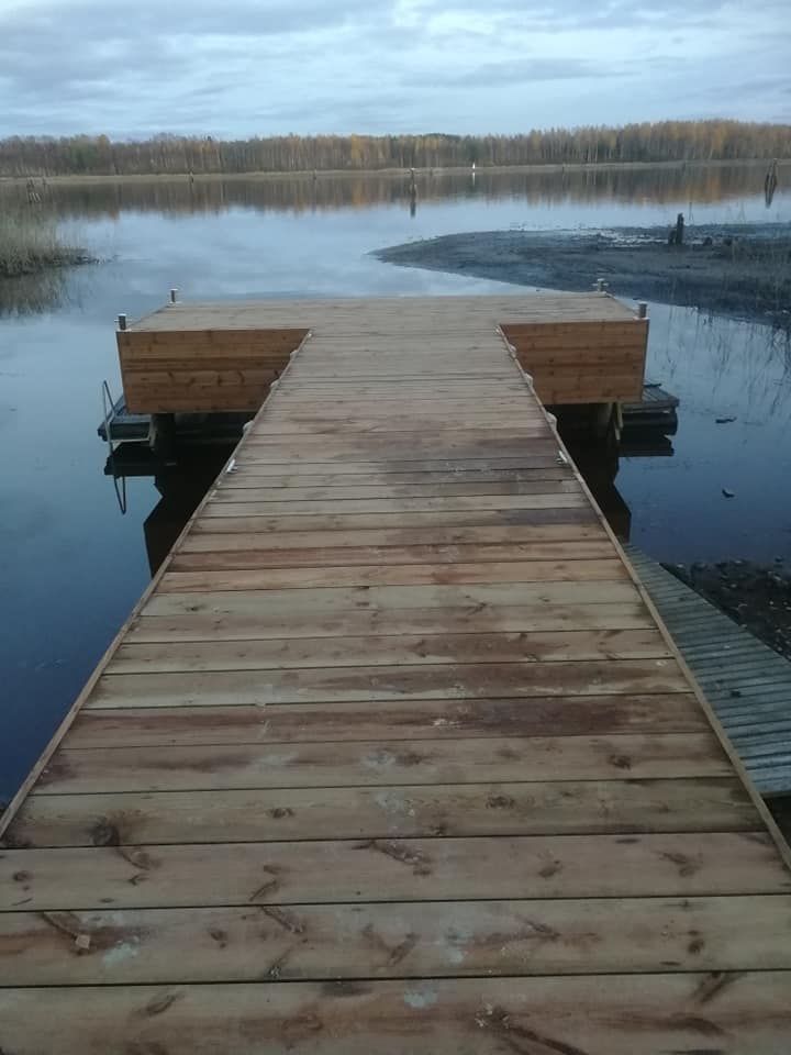 Puinen laituri ulottuu tyynen järven ylle, taustalla metsä ja pilvinen taivas.