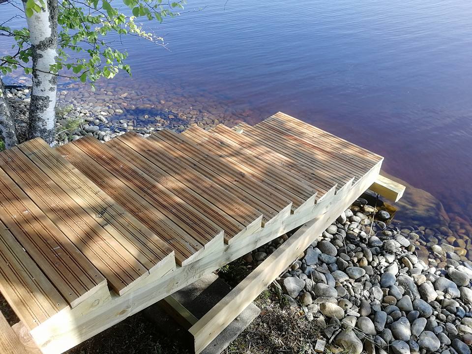 Puinen laituri laskeutuu kivikkoiselle rannalle, vieressä koivu ja tumma vesi.