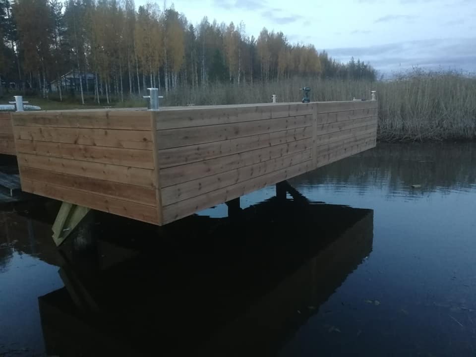 Puinen laituri seisoo veden päällä, taustalla puita ja ruokoja, hämärä valaistus.