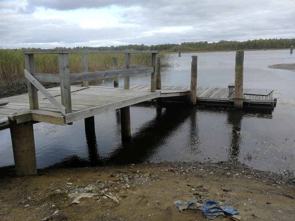 Kuvassa on puinen laituri, joka ulottuu tummaan veteen. Taustalla näkyy kaislikkoa ja puita pilvisen taivaan alla. Rannalla on hiekkaa ja roskia.