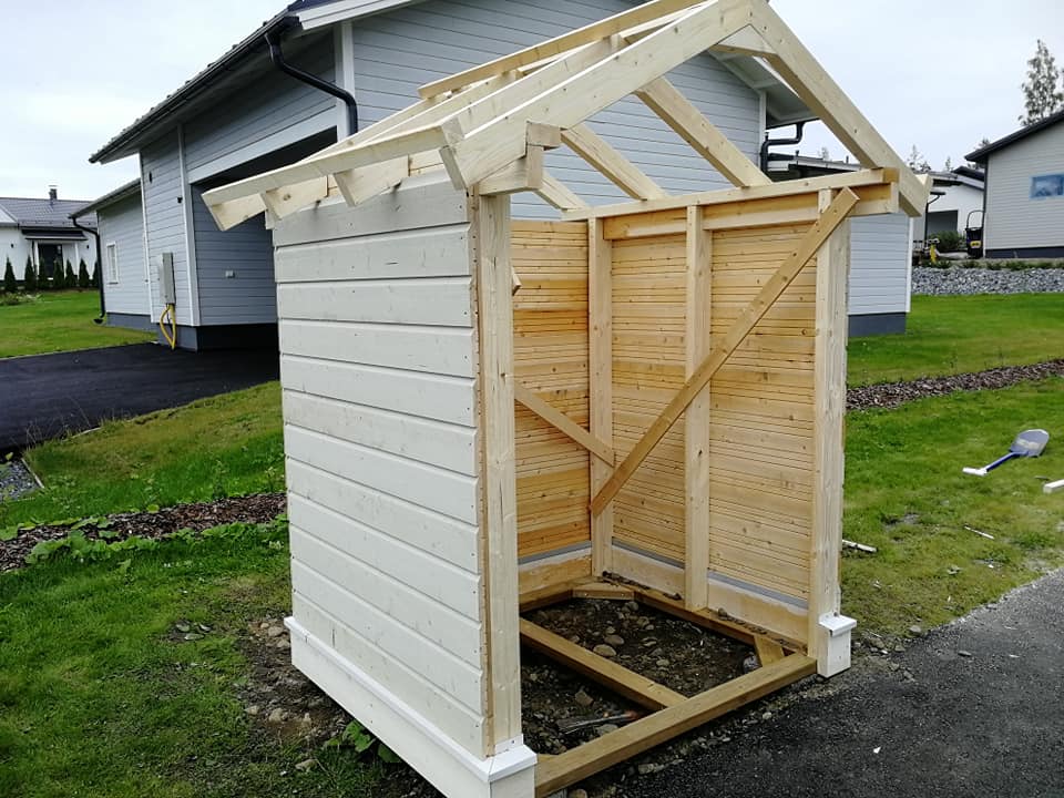 Keskeneräinen puurakennelma, jossa on vaaleat seinät ja puinen katto, sijaitsee nurmikolla valoisassa säässä.