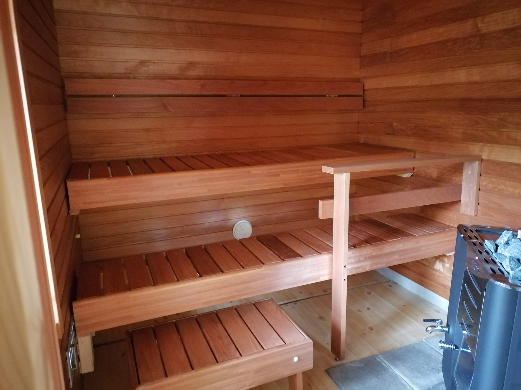 Puinen sauna, jossa on kaksi penkkiä, pieni jakkara ja kiuas, valaistus on lämmin.