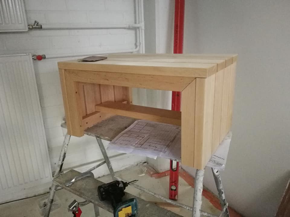 Puinen sivupöytä rakennustelineillä, jossa on porakone, suunnitelmat ja hiomapaperi, valaistus on kirkas.