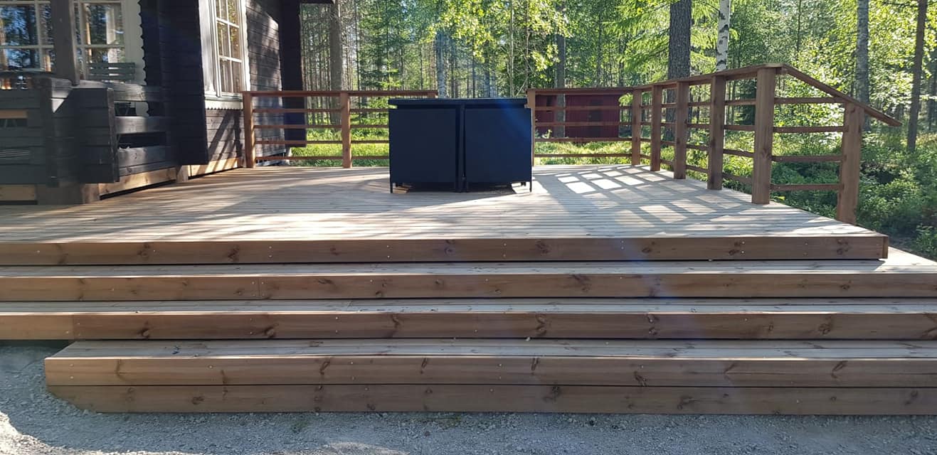 Ulkoterassi, jossa on puuportaat, puukaide ja musta säilytysyksikkö, valaistu auringonvalolla.