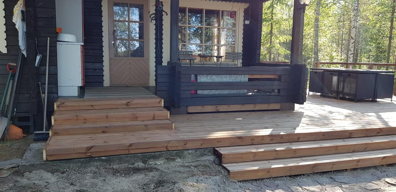 Kuva esittää puisen terassin, jossa on ruskeat portaat ja musta kaide. Terassilla on myös musta ulkokaluste. Valoisa sää.