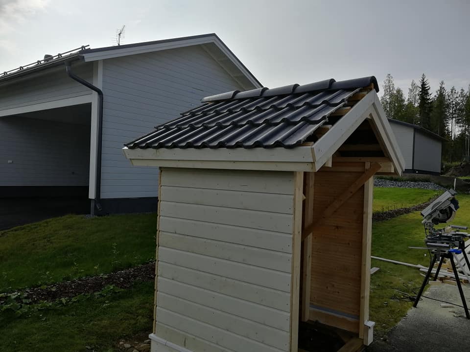 Pieni puinen ulkorakennus, jossa on musta tiilikatto, sijaitsee nurmikolla harmaan talon ja sirkkelin lähellä.