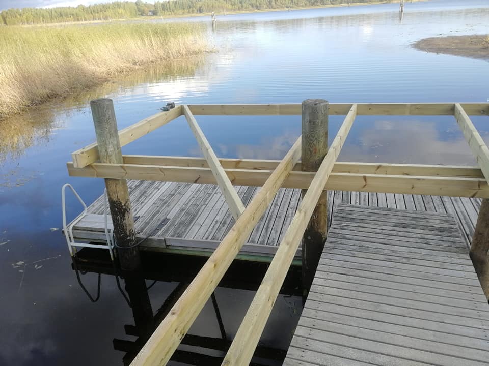 Kuva esittää puista laituria järvellä, jossa on uusi puurunko rakenteilla. Laiturissa on harmaita puulankkuja ja valkoiset portaat. Järven vesi on tyyni ja heijastaa taivasta.