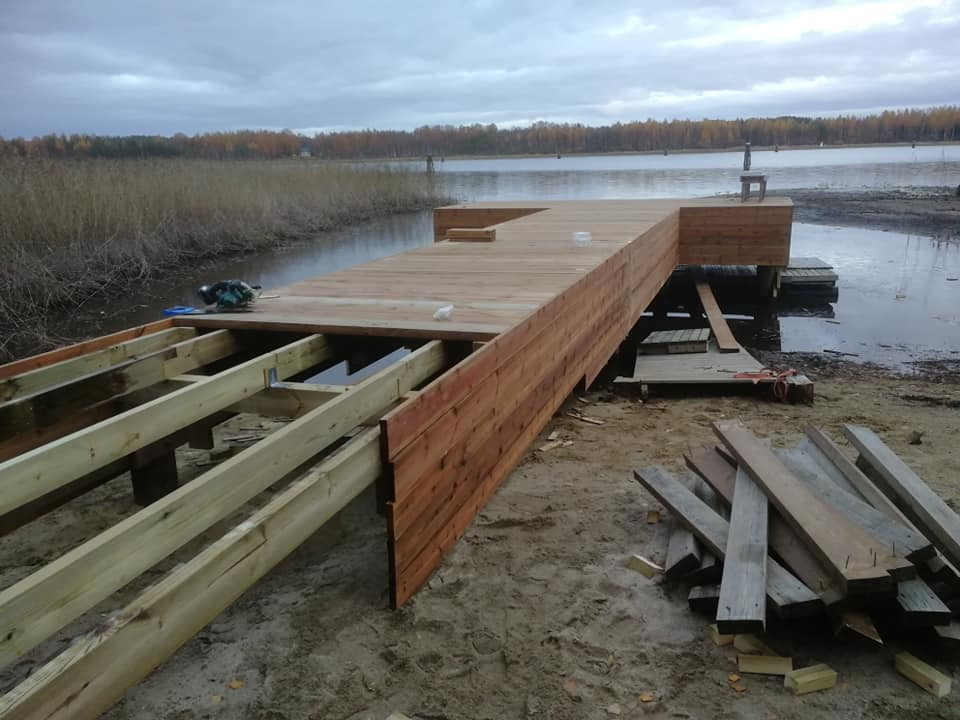 Kuva esittää rakenteilla olevaa puista laituria järven rannalla, jossa näkyy osittain valmiita osia ja kasaamattomia puutavaroita.