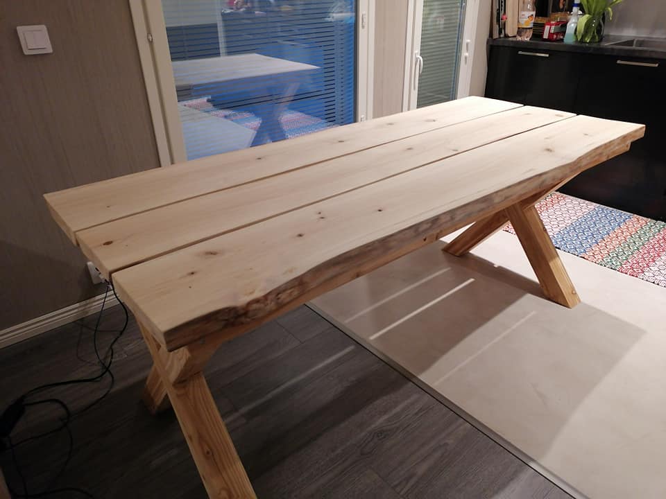 Vaalea puinen ruokapöytä, jossa on X-muotoiset jalat, seisoo vaalealla matolla harmaiden lattialautojen päällä. Pöydän takana on ikkuna ja keittiön työtaso.