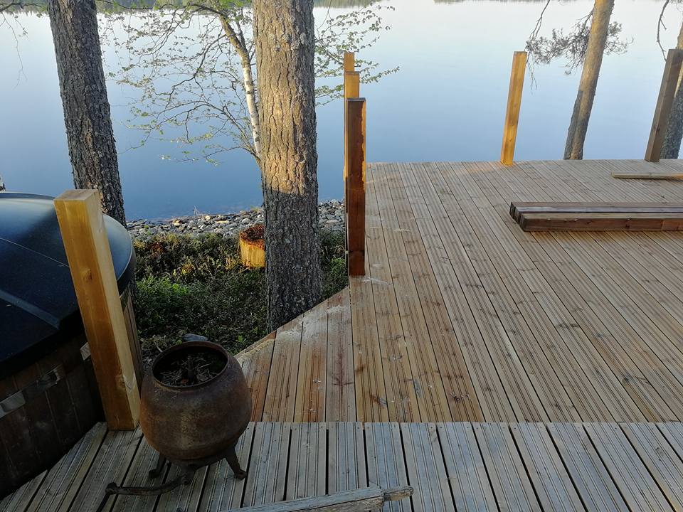 Ulkona on puinen terassi, jolla on puita, järvi taustalla ja ruosteinen pata etualalla. Valo on pehmeää ja luonnollista.