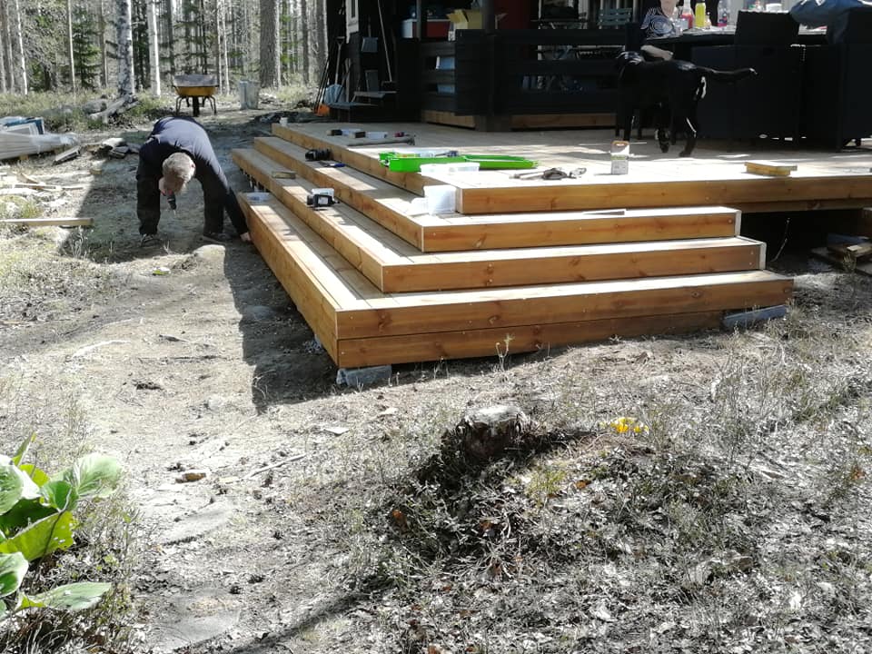 Ulkona on rakenteilla puinen terassi, jossa on neljä porrasta. Mies työskentelee terassin vieressä. Taustalla näkyy metsää ja ihmisiä.