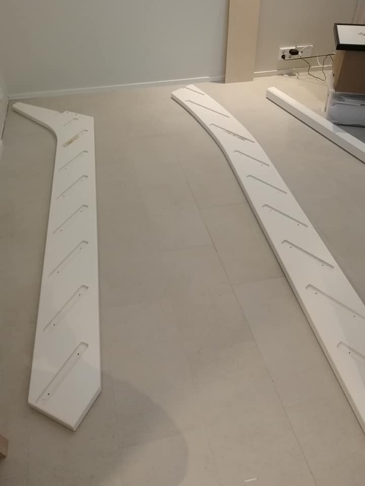 Kaksi valkoista, kaarevaa puukappaletta lattialla, joissa on viivoja ja reikiä, sekä valkoinen lista ja pahvilaatikko taustalla.