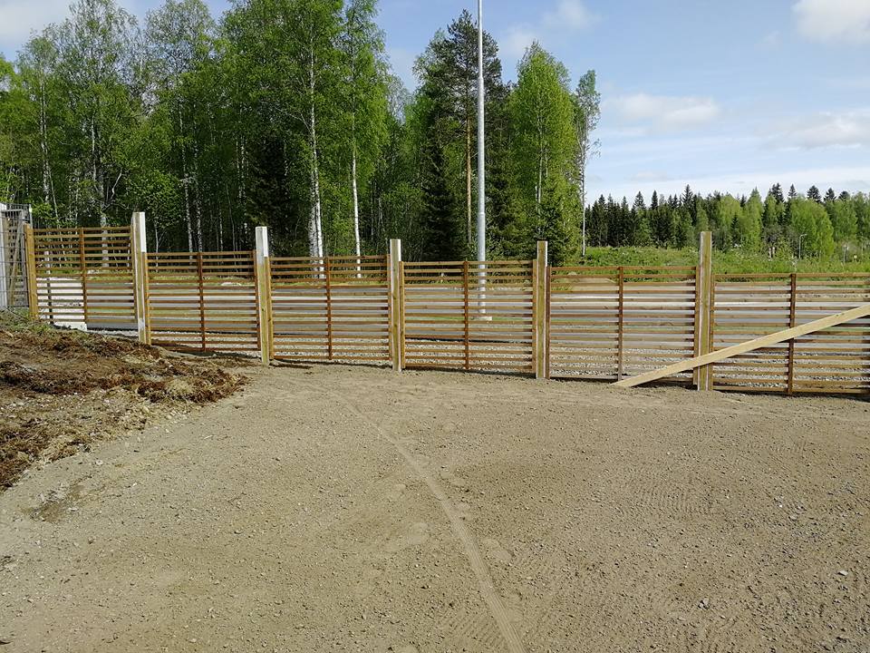 Vaalea puuaita hiekkakentällä, taustalla metsä ja sininen taivas.