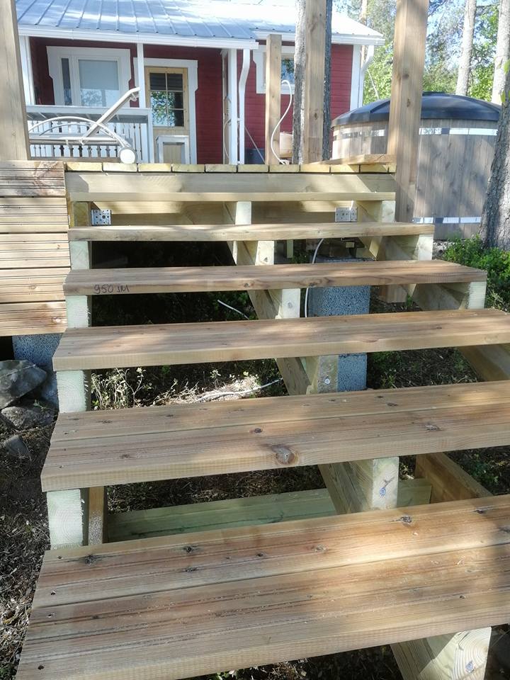 Ulkona on puuportaat, jotka johtavat punaiseen taloon ja puiseen kylpytynnyriin. Portaat on rakennettu vaaleasta puusta ja ne ovat osittain varjossa.