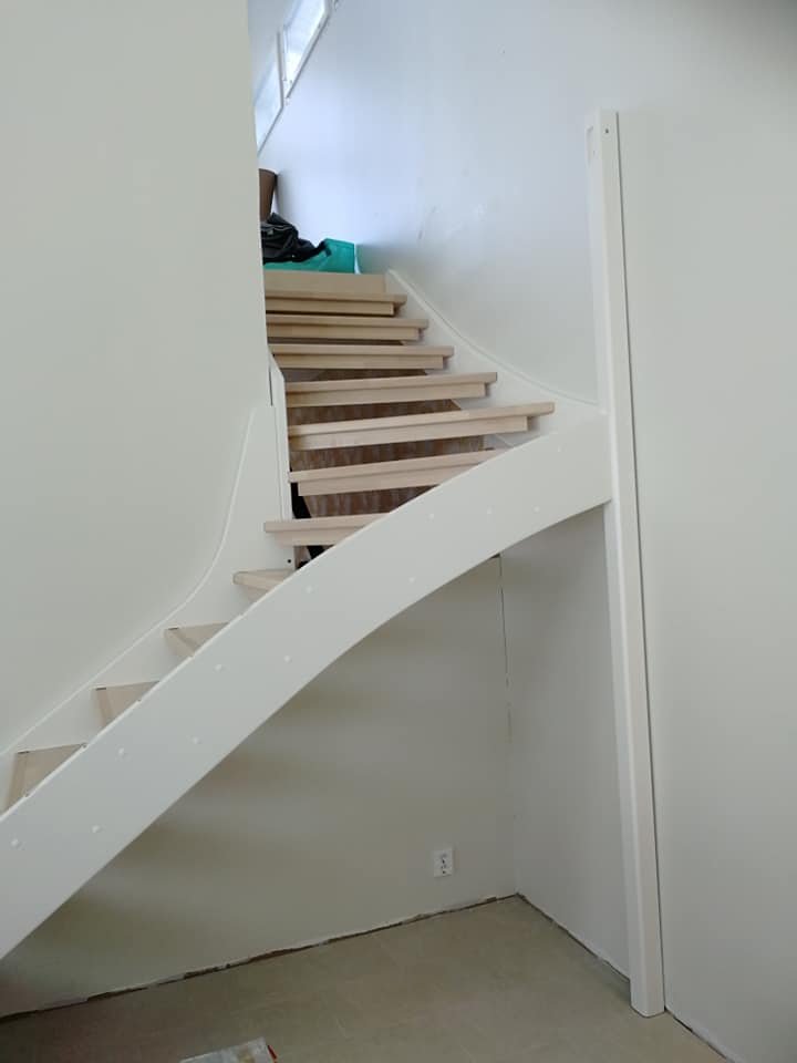 Vaaleat puuportaat valkoisella kaiteella valkoisessa huoneessa, jossa on ikkuna ylhäällä ja pistorasia seinässä.