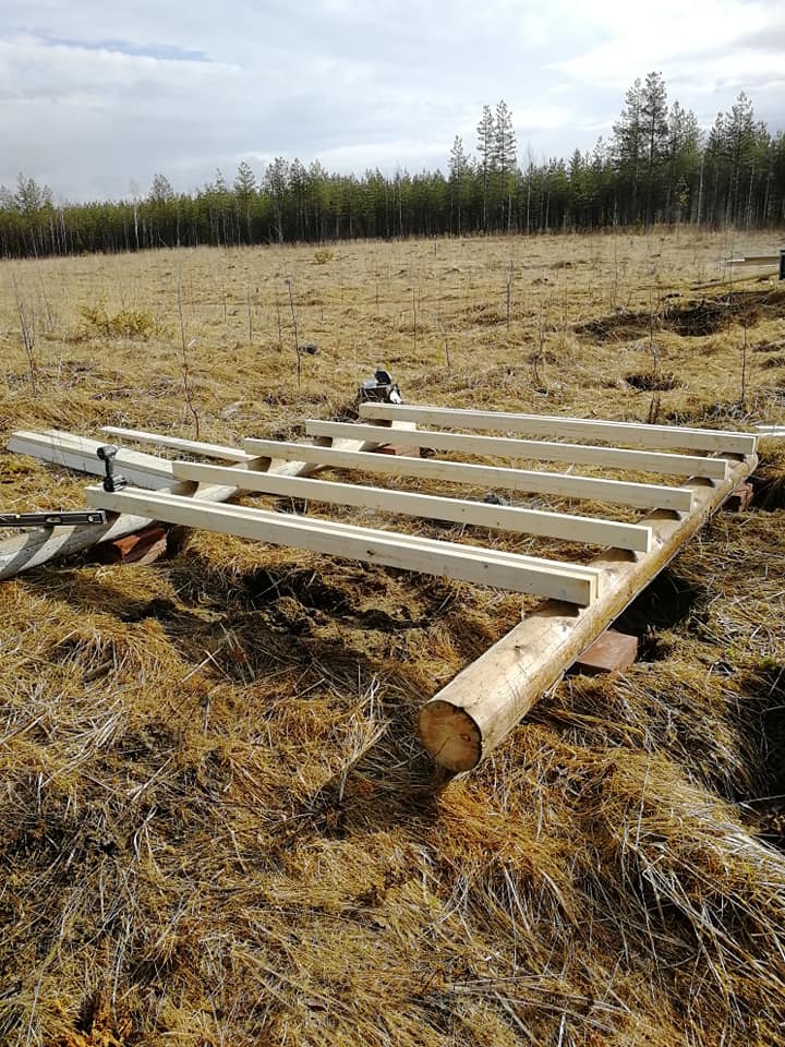 Maaseudulla oleva puurakennelma, jossa on tukipuu ja useita vaakasuoria lautoja, jotka lepäävät tiilien päällä, kuivassa ruohikossa, taustalla metsä ja pilvinen taivas.