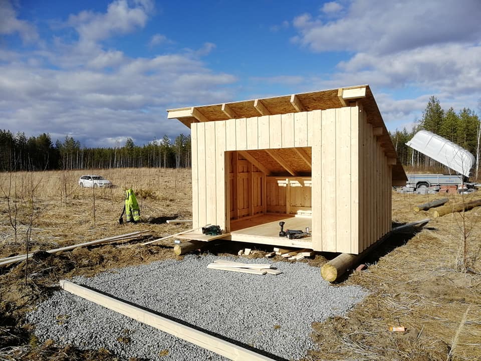 Keskeneräinen puurakennus maaseudulla, jossa on harmaa kivipohja ja sininen taivas.