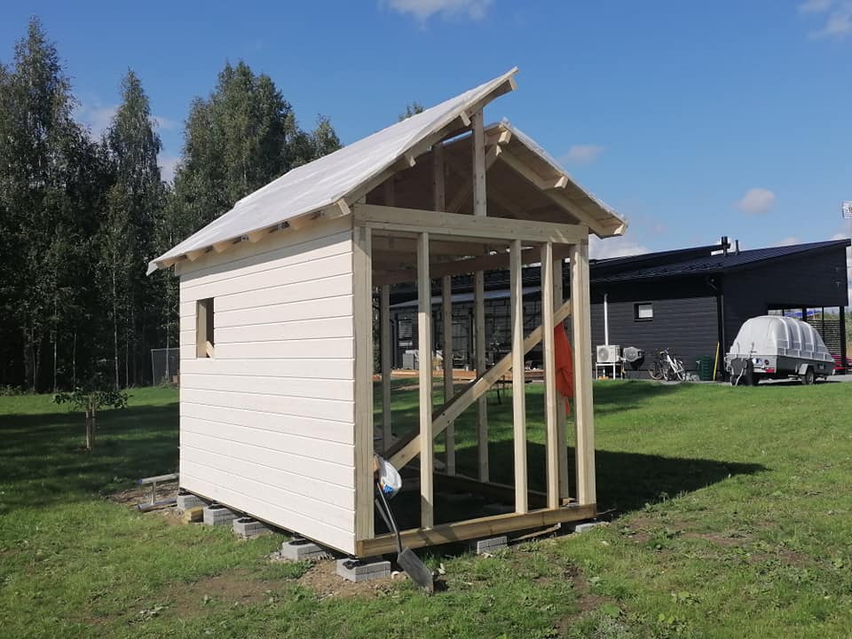 Keskeneräinen puurakenteinen rakennus, jossa on valkoiset seinät ja läpinäkyvä katto, seisoo vihreällä nurmikolla aurinkoisena päivänä.