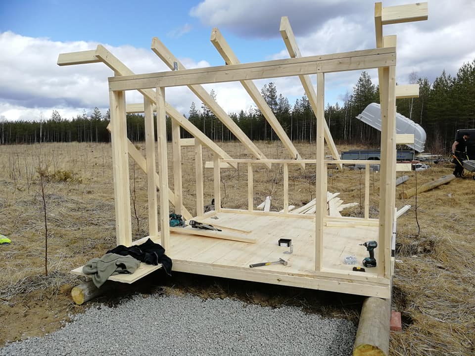 Keskeneräinen puurakenteinen rakennus, jossa on puinen lattia, pystypuut ja kattopalkit, ulkona maaseudulla aurinkoisena päivänä.