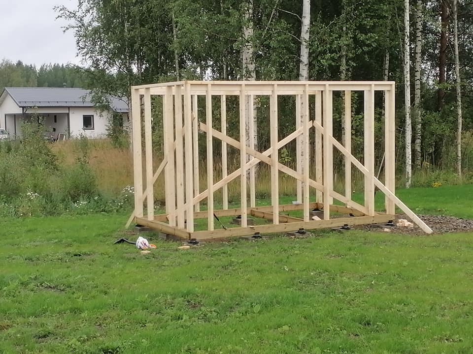 Keskeneräinen puurunkoinen rakennus vihreällä nurmikolla, taustalla talo ja puita, valoisa sää.