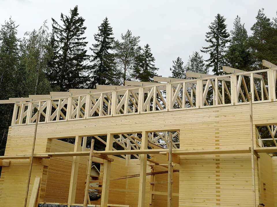 Rakenteilla oleva puutalo, jossa näkyy puurakenteet ja taustalla metsä. Vaalea puu ja harmaa taivas.