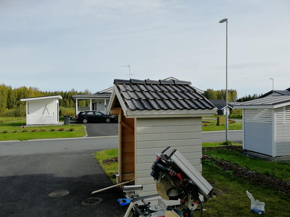 Kuvassa näkyy vaalea puuvaja, jonka katto on tummanharmaa. Etualalla on sahauspöytä ja taustalla näkyy asuinalue, jossa on muita rakennuksia ja auto. Taivas on vaalea ja valaistus on luonnollinen.