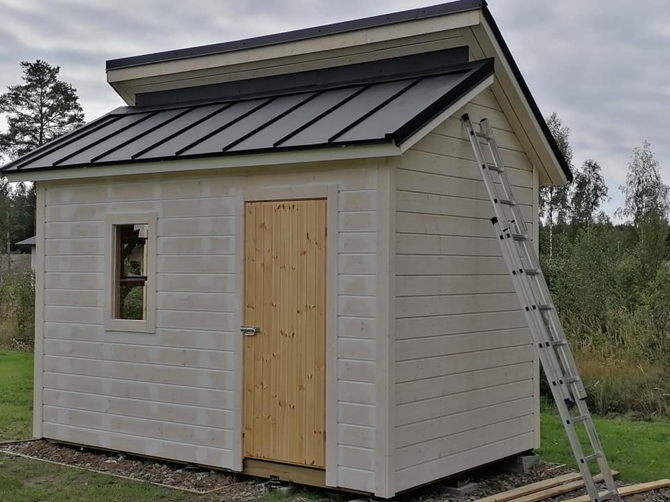 Pieni, valkoinen puuvaja, jossa on harmaa peltikatto ja puinen ovi, seisoo nurmikolla, ja sen vieressä on alumiininen tikapuu.