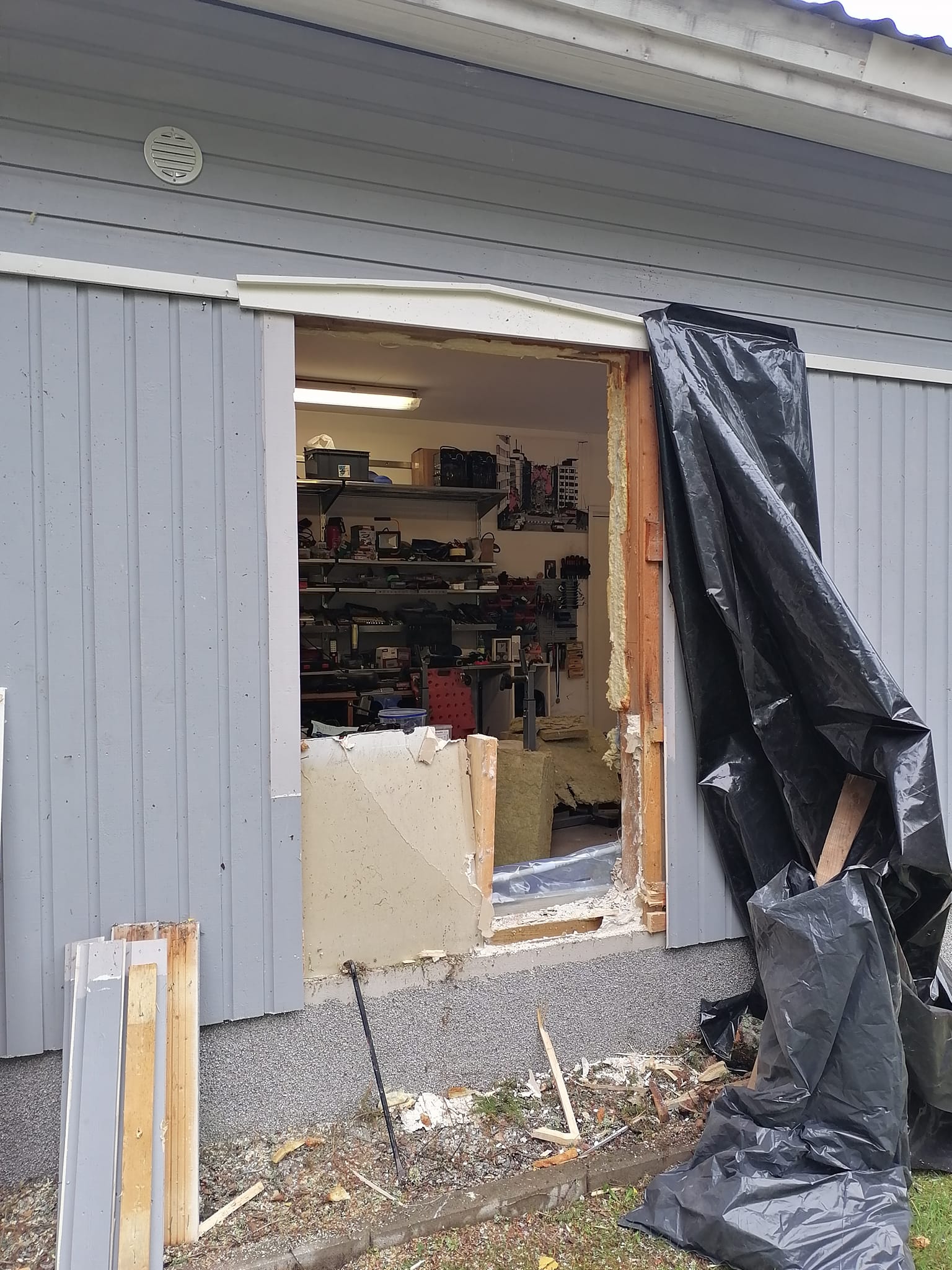 Ulkokuva rakennuksesta, jossa on avoin aukko seinässä, peitettynä mustalla muovilla. Aukon sisällä näkyy työkaluja ja hyllyjä. Rakennuksen seinät ovat vaaleanharmaat, ja maassa on rakennusjätettä.
