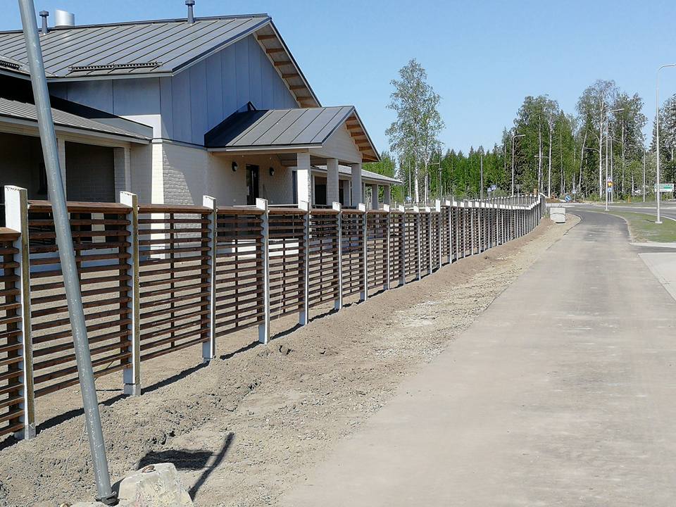 Vaalea rakennus ruskealla aidalla, valaistus on kirkas ja aurinkoinen.