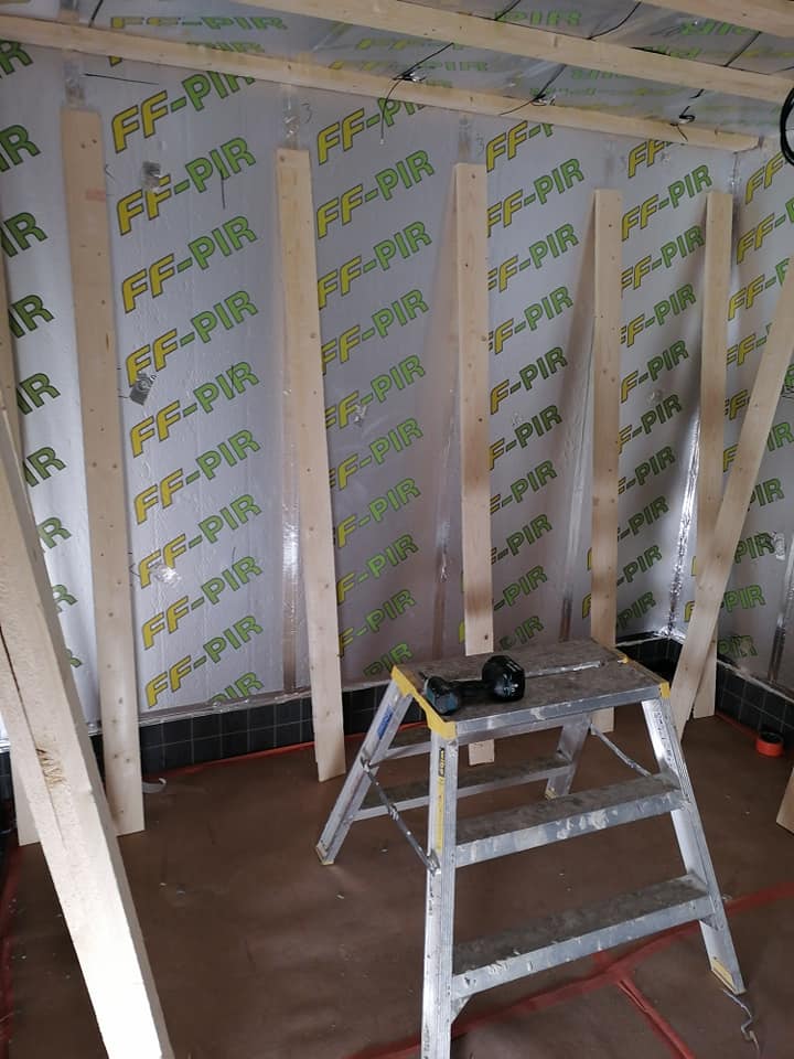 Rakennustyömaa, jossa on FF-PIR-eristelevyillä vuorattu seinä, pystytolpat ja alumiiniset tikkaat, joilla on porakone. Valoisa sisätila.